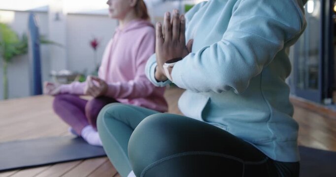 Diverse women plus senior settling on balcony mats raising hands in mudra for calm showing watch