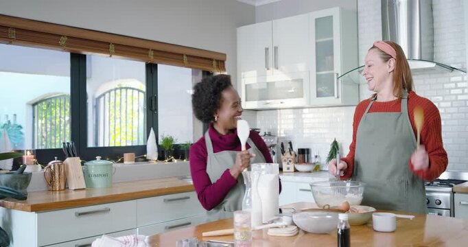 Diverse female friends checking phone for recipe, whisking batter and miming with spatula at island