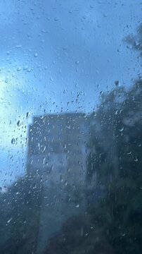 Relaxing visual loop of blue-tinted raindrops hitting a window. A blurred city building outside creates an urban aesthetic, perfect for modern motion graphics, social media, or calm weather clips.