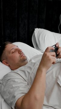 Close-up of a person's hands holding a black mobile phone in a horizontal position while resting in a cozy bed. Morning smartphone use, scrolling social media, or mobile gaming concept with selective