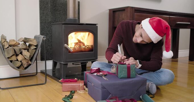 African American man in Santa hat sitting beside lit wood stove wrapping and tagging holiday gifts