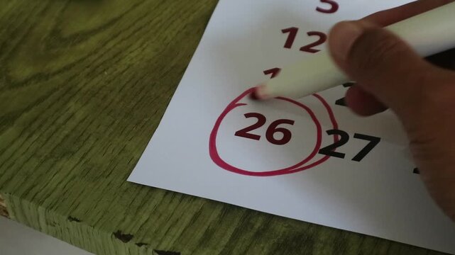 A hand using a red marker to circle the twenty-sixth day on a wall calendar for vacation travelling during holiday