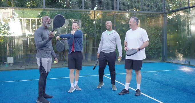 Diverse male friends on blue paddle court with paddles, man bouncing ball, preparing casual match