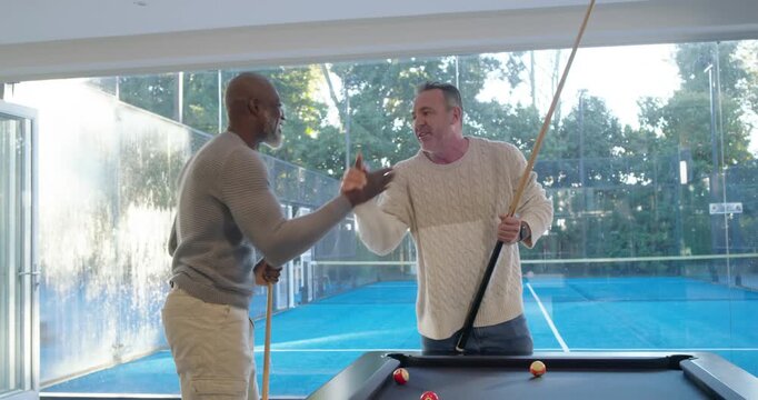 Two men one African American alternating shots at pool table after shake glass showing tennis court