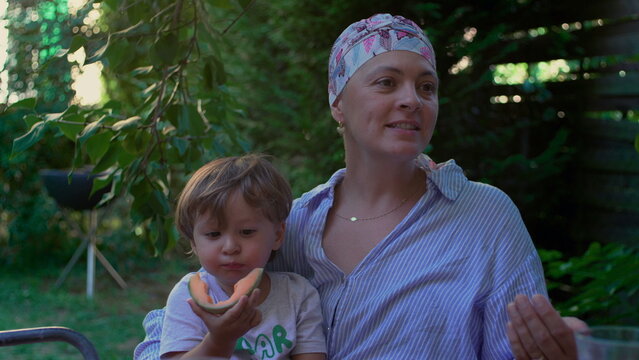 Mother with cancer holding toddler eating melon in a garden, nurturing outdoor family moment expressing resilience, connection and everyday joy through illness