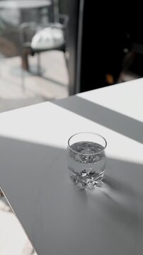 Close-up of a clear glass filled with fresh drinking water standing on a tabletop near a window with harsh sunlight shadows. Concept of healthy lifestyle, hydration, purity and morning routine.