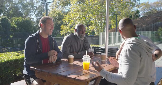 Diverse men reacting to gray hoodie man entering, sitting at patio table, toasting cups for meetup