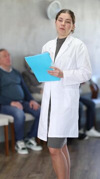 Portrait of positive young female secretary standing with folder with documents and materials in hands and inviting in cabinet or to appointment