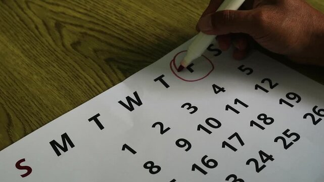 A hand using a red marker to circle a date on a paper calendar.