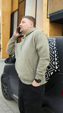 Millennial male holding mobile phone and having a conversation outdoors next to an automobile. Concept of modern urban communication, cellular connection, business talk, or calling a taxi.