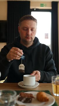 Close-up of a millennial male traveler dipping a tea bag into a white ceramic mug at a table with a croissant and pastry. Morning routine, breakfast time, and cozy restaurant lifestyle concept.