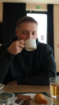 Close-up of a pensive millennial male traveler holding a white ceramic mug and taking a sip of hot beverage at a restaurant table. Morning routine, breakfast time, and coffee break concept.