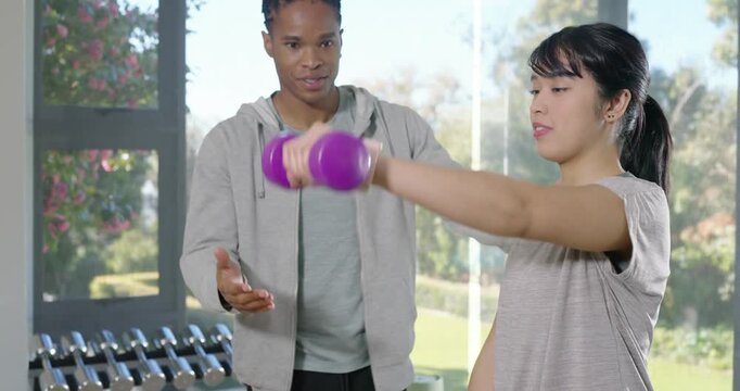 Male trainer guiding Asian trainee doing front raises with purple dumbbell in gym, correcting form