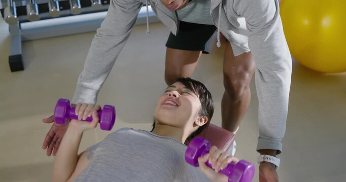 African American trainer spotting Asian client pressing purple dumbbells on incline bench for form