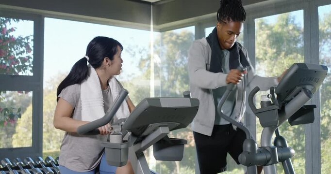 African American man and Asian woman pedaling at gym on bike, elliptical, woman asking, man turning