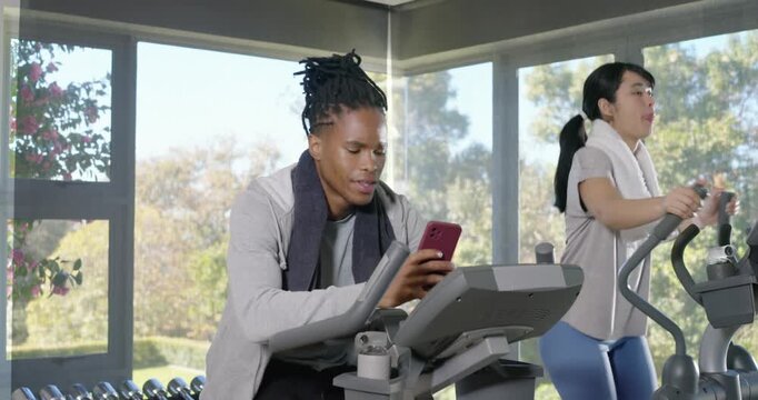 Diverse friends pedaling in bright gym room, man retrieving pink smartphone, sharing funny message