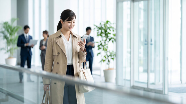 オフィスビルのロビーでスマートフォン操作をするトレンチコート姿のビジネスパーソン女性