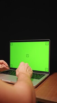 Vertical video. Hands Type on a Laptop Keyboard With Green Screen Display on Wooden Desk, Supporting Digital Technology and Screen Replacement