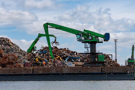 Green material handler cranes operating at port scrap metal recycling yard with huge metal piles under cloudy sky, circular economy concept