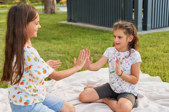 Girls sit facing each other on a blanket in a park. They are smiling and playing a hand clapping game. Nearby, trees and a building are visible