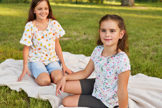 Two girls spend time on a blanket in a park. They have fun together under the sun in the afternoon. The grass is green around them. They smile and chat