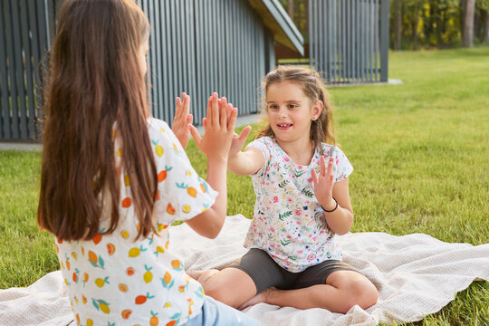 Two girls are sitting on a blanket in a grassy area. They are playing a hand game with smiles. Trees are visible in the background under a clear sky