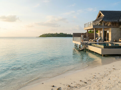 A woman stands on the balcony of an overwater bungalow overlooking a turquoise ocean at sunset