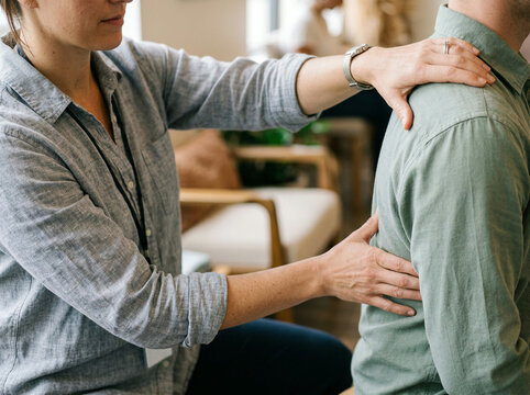 A woman places her hands on a man's back and shoulder in a supportive gesture indoors