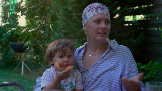 Mother with cancer holding toddler eating melon in a garden, nurturing outdoor family moment expressing resilience, connection and everyday joy through illness