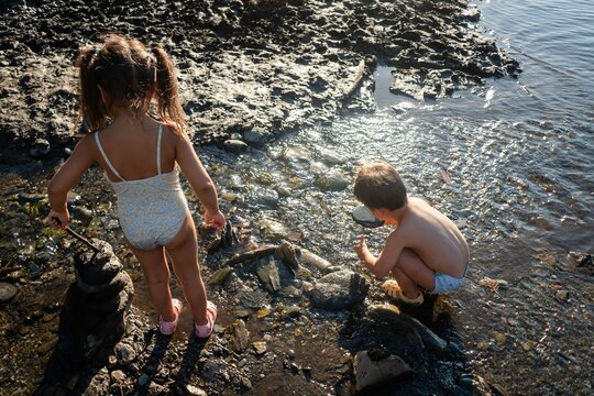 Two white children play in mud and shallow water along a riverbank
