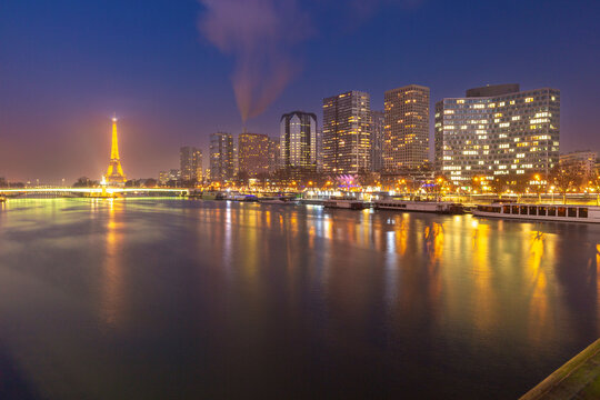Night view across the Seine in Paris, France, with modern high-rise buildings, riverboats and city lights reflected on the water.