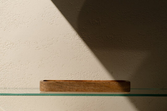 Small empty wooden tray placed on a transparent glass shelf in front of a travertine effect plaster wall with a dramatic diagonal shadow, serving as a minimal cosmetic product staging prop.