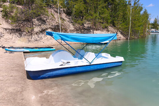 White pedal boat with blue canopy moored at a sandy lakeshore, surrounded by forest and calm green water on a sunny summer day. Ideal for travel, vacation and outdoor recreation concepts.