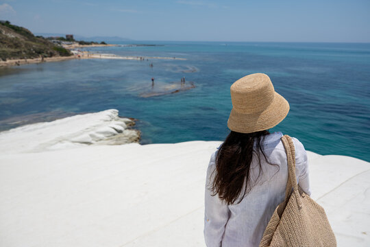 Woman in straw hat standing on white rock and looking at blue sea coastline in Sicily