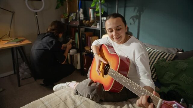 Young Caucasian woman playing acoustic guitar while sitting on bed, camera zooming out. Footage is perfect for promoting music lessons, creative hobbies, or online learning platforms