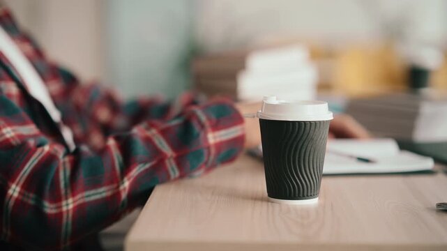Student in plaid shirt studies at wooden desk with laptop and coffee cup