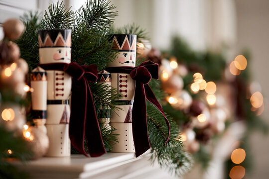 Decorative nutcracker figurines with red bows positioned on a mantelpiece adorned with evergreen garlands and warm glowing lights for a festive holiday atmosphere