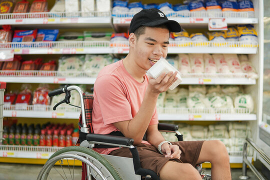 Young Asian man in wheelchair holding food product package while grocery shopping in supermarket. Disabled male shopper enjoying independent lifestyle, inclusion and accessibility concept.