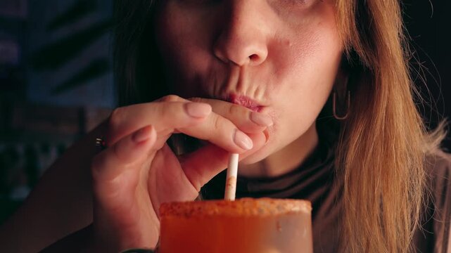 Close-up of a young woman sipping a spicy michelada cocktail through a straw in a restaurant. Refreshing beer-based drink with chili rim, casual dining, and social gathering concept. High quality 4k