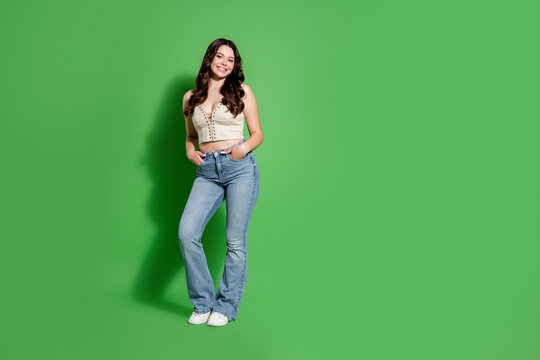 Smiling young woman in a casual crop top and jeans standing against a green background, full-length portrait conveying confidence and relaxed lifestyle