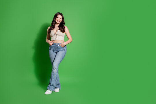 Smiling young woman in casual jeans and crop top standing in a studio with a green background and relaxed confident pose for lifestyle and fashion portraits