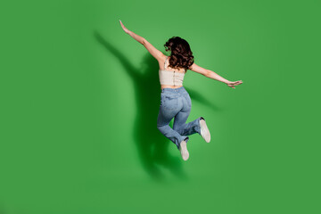 Fototapeta premium Woman jumping midair in jeans on a green background expressing joy, motion and carefree energy in a studio fashion lifestyle pose