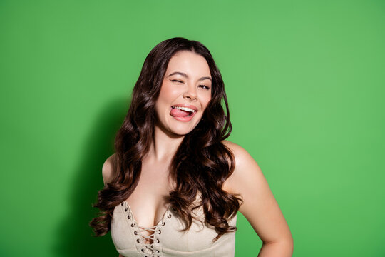 Playful young woman with long brunette hair winking and sticking out her tongue against a green studio backdrop, casual fashion portrait conveying fun, confidence and flirtatious charm