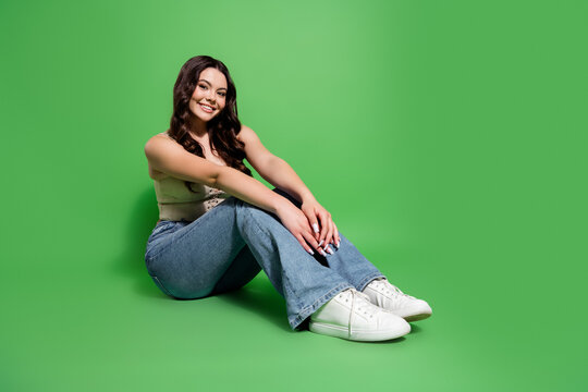Smiling young woman sitting on green background in jeans and white sneakers for casual studio portrait and lifestyle fashion