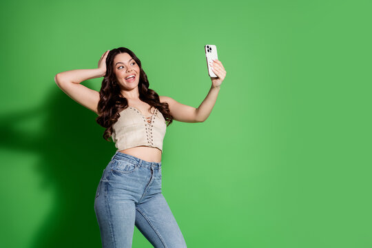 Happy young woman taking a selfie with smartphone against green background, casual crop top and jeans, influencer-style portrait showing joy and confidence