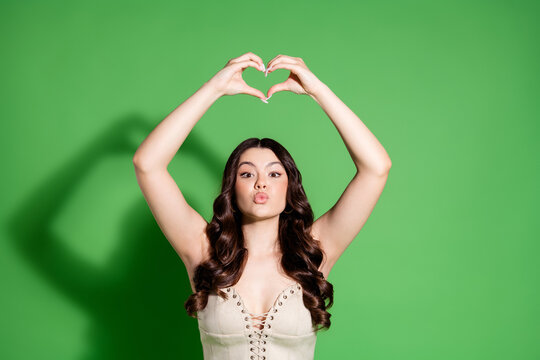 Young woman making a heart gesture above her head and blowing a kiss against a green studio background