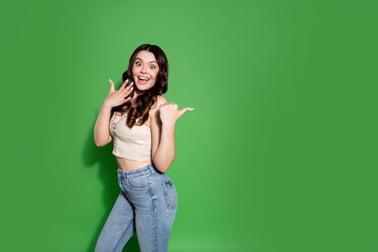 Happy young woman pointing and smiling over her shoulder on green background, casual portrait of a surprised brunette in crop top and jeans