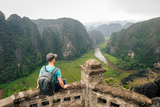Tourist with backpack sitting on viewpoint, enjoying scenic view of rice fields and river amidst karst mountains, Ninh Binh Vietnam. Southeast Asia travel, outdoor exploration and wanderlust concept.