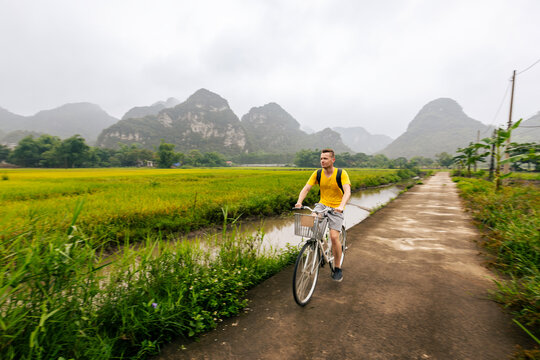Tourist riding bicycle on road between rice fields with limestone karst mountains background in Ninh Binh Vietnam. Eco tourism, outdoor cycling adventure and rural Asian travel concept.