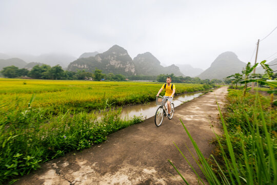 Tourist riding bicycle on road between rice fields with limestone karst mountains background in Ninh Binh Vietnam. Eco tourism, outdoor cycling adventure and rural Asian travel concept.